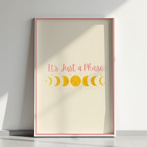 Puede incluir: Un estampado enmarcado con un borde rosa claro. La obra de arte presenta la frase "It's Just a Phase" en escritura rosa sobre una serie de fases lunares amarillas. El fondo es de color crema suave.
