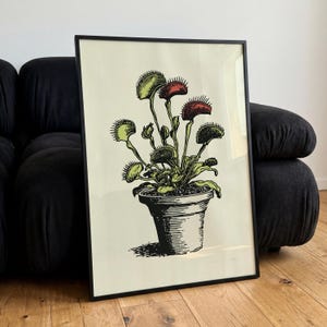 May include: A framed print featuring a black ink illustration of a Venus flytrap plant in a pot. The plant has green leaves and red-tinted traps with spiky edges. The background of the print is a light cream color, and the frame is thin and black.