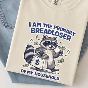 May include: Cream-colored sweatshirt with the text "I AM THE PRIMARY BREADLOSER OF MY HOUSEHOLD." A cartoon raccoon wearing sunglasses and holding a martini and a shopping bag filled with money is printed on the front. The sweatshirt is laid on top of blue jeans.