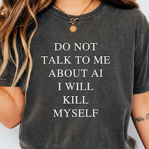 May include: A charcoal grey short-sleeved t-shirt featuring the white text DO NOT TALK TO ME ABOUT AI I WILL KILL MYSELF printed in a clean, sans-serif font across the center of the chest. The garment has a crew neckline and a relaxed fit.