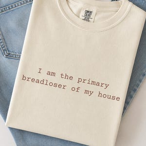 May include: Cream-colored crew neck sweatshirt with the text "I am the primary breadloser of my house" in brown lettering. The sweatshirt is laid on top of a pair of light blue denim jeans.