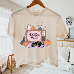 May include: A cream-colored t-shirt featuring a graphic of a vintage television set with the text "PRACTICAL MAGIC" on the screen. The design includes a black cat, two orange pumpkins, a broomstick, and two candles on a purple rug.