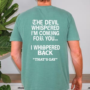 May include: A sage green short-sleeve t-shirt featuring white text on the back that reads: THE DEVIL WHISPERED I'M COMING FOR YOU... I WHISPERED BACK "THAT'S GAY". The lettering uses a mix of gothic-style and standard sans-serif fonts.