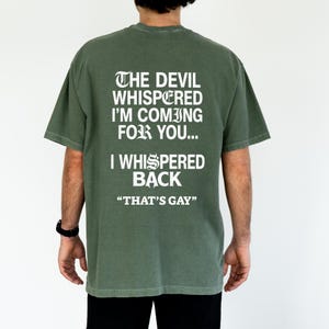 May include: An olive green short-sleeved t-shirt featuring white text on the back that reads: THE DEVIL WHISPERED I'M COMING FOR YOU... I WHISPERED BACK "THAT'S GAY". The shirt has a crew neckline and a relaxed fit, displayed from the back view.
