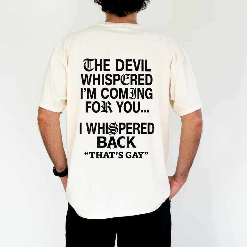 May include: An off-white short-sleeve t-shirt featuring black text on the back that reads: THE DEVIL WHISPERED I'M COMING FOR YOU... I WHISPERED BACK "THAT'S GAY". The lettering is in a bold, gothic-style font with a distinct, stylized letter S.