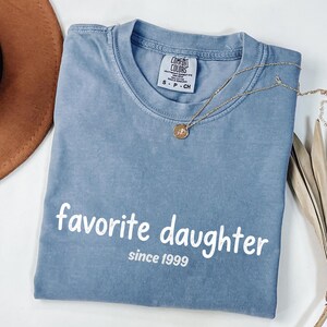 May include: A blue Comfort Colors t-shirt with the words "favorite daughter since 1999" printed in white. A gold necklace with a pendant is draped across the shirt.