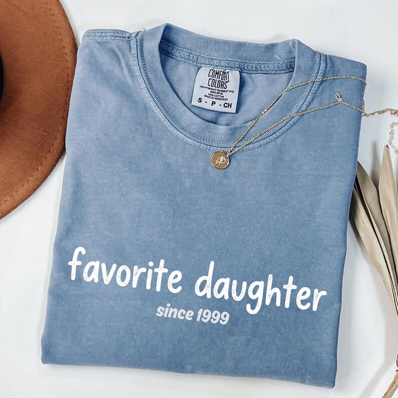 May include: A blue Comfort Colors t-shirt with the words "favorite daughter since 1999" printed in white. A gold necklace with a pendant is draped across the shirt.