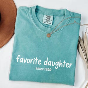 May include: A teal Comfort Colors t-shirt with the words "favorite daughter since 1999" in white. A gold necklace with a pendant rests on the shirt. A brown hat is partially visible in the upper left corner.