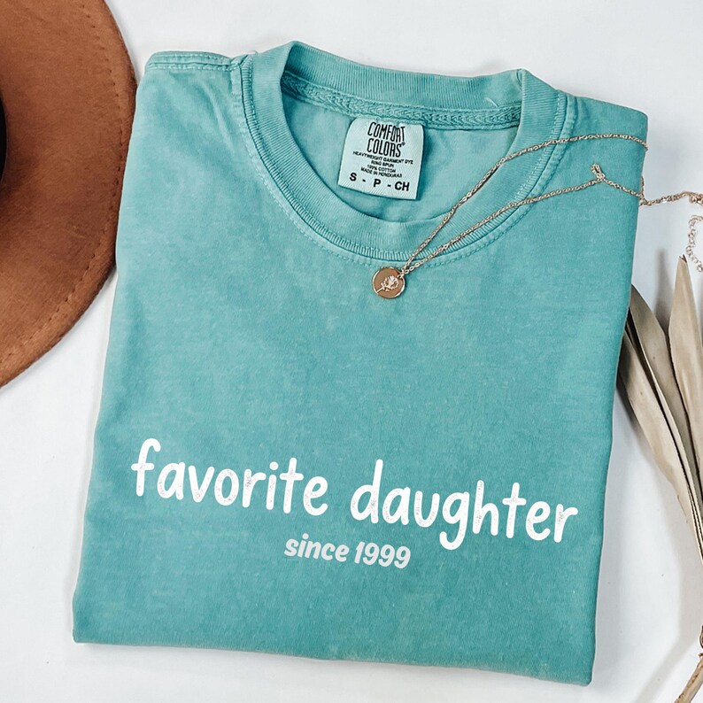 May include: A teal Comfort Colors t-shirt with the words "favorite daughter since 1999" in white. A gold necklace with a pendant rests on the shirt. A brown hat is partially visible in the upper left corner.