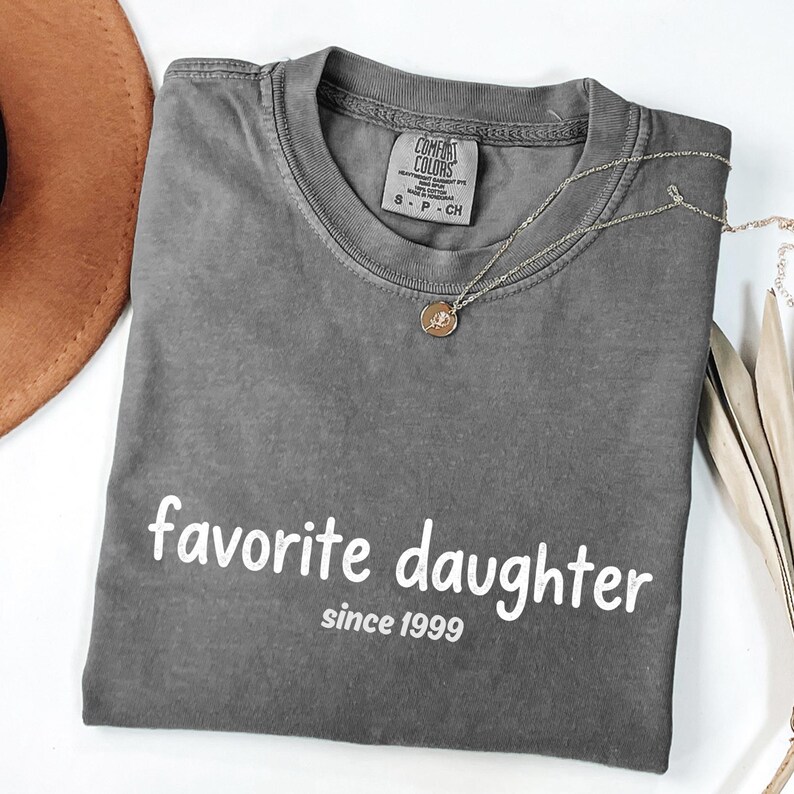 May include: A grey Comfort Colors t-shirt with the words "favorite daughter since 1999" printed in white. A silver necklace with a small pendant is draped across the shirt.