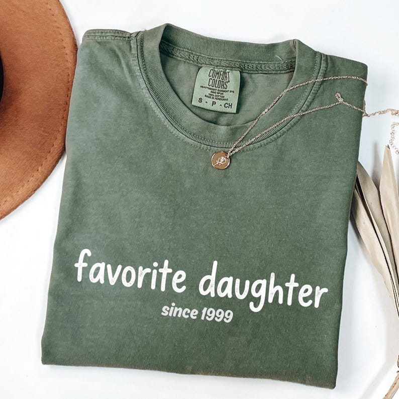 May include: An olive green t-shirt with the words "favorite daughter" and "since 1999" printed in white. A gold necklace with a pendant is draped across the shirt. A brown hat is visible in the upper left corner.