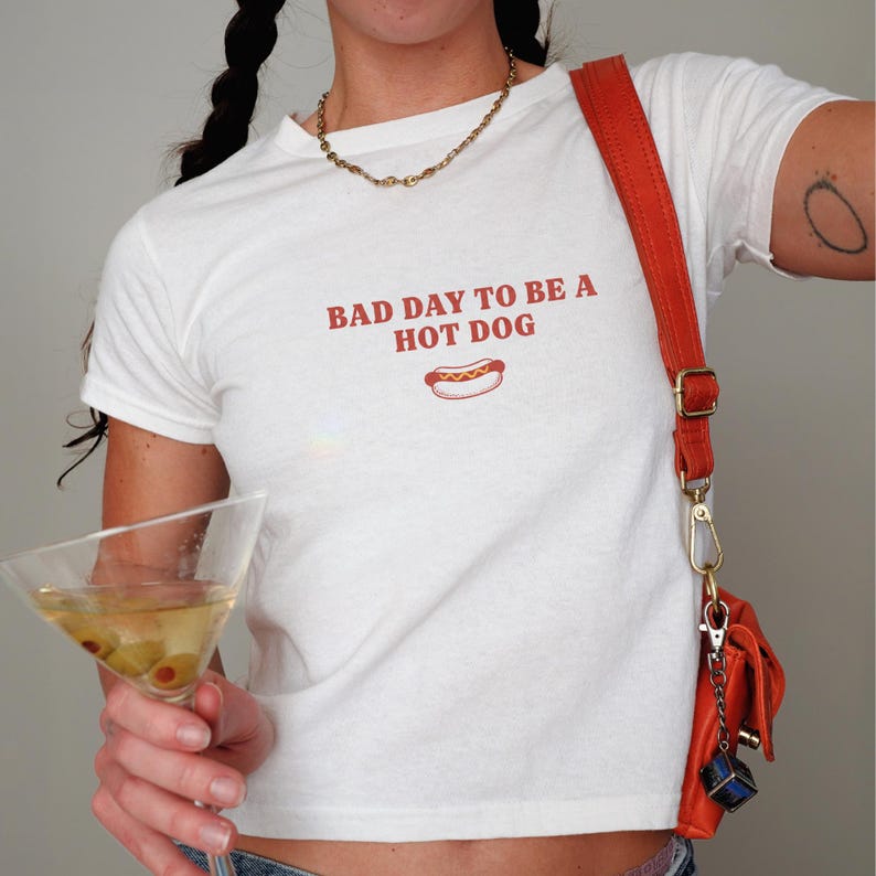 以下が含まれることがあります： 「BAD DAY TO BE A HOT DOG」という赤い文字とホットドッグの小さなグラフィックがプリントされた白い半袖Tシャツ。ゴールドのチェーンネックレスと、小さな青いキューブのチャームが付いたオレンジ色のクロスボディバッグを合わせています。