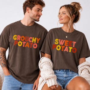 May include: Two dark brown short-sleeve t-shirts. The left shirt features the text "GROUCHY POTATO" in distressed yellow, orange, and red lettering. The right shirt features the text "SWEET POTATO" in the same distressed yellow, orange, and red colors.