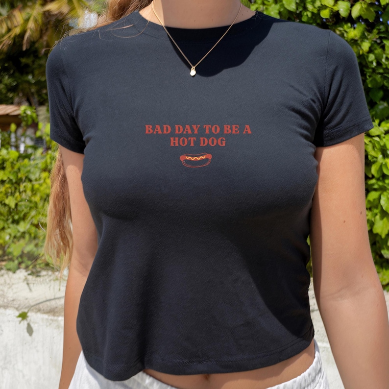 以下が含まれることがあります： 胸元に赤い文字で「BAD DAY TO BE A HOT DOG」とプリントされ、黄色いマスタードがかかったホットドッグの小さなグラフィックイラストが添えられた、黒い半袖Tシャツ。