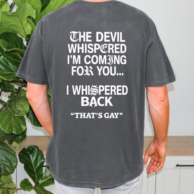 May include: A charcoal gray short-sleeve t-shirt featuring white text on the back that reads: THE DEVIL WHISPERED I'M COMING FOR YOU... I WHISPERED BACK "THAT'S GAY". The shirt has a crew neckline and a relaxed fit, displaying the back side only.