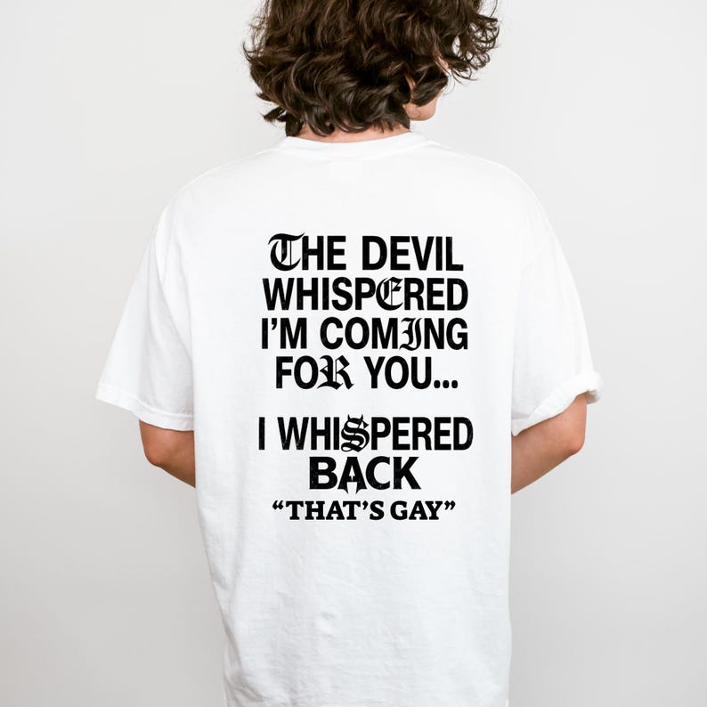 May include: A white short-sleeved t-shirt featuring black text on the back that reads: THE DEVIL WHISPERED I'M COMING FOR YOU... I WHISPERED BACK "THAT'S GAY". The lettering is rendered in a bold, gothic-style font with stylized characters.