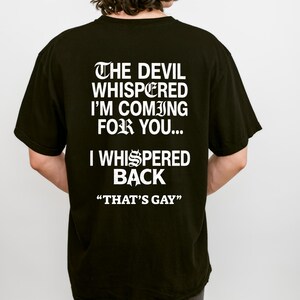 May include: A black short-sleeved t-shirt featuring white text on the back that reads: THE DEVIL WHISPERED I'M COMING FOR YOU... I WHISPERED BACK "THAT'S GAY". The lettering uses a mix of gothic-style and standard sans-serif fonts.