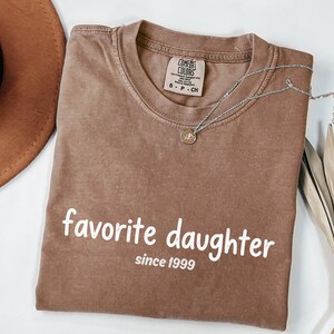 May include: A brown Comfort Colors t-shirt with the words "favorite daughter since 1999" printed in white. A silver necklace with a small pendant is draped across the shirt. The shirt is folded neatly.