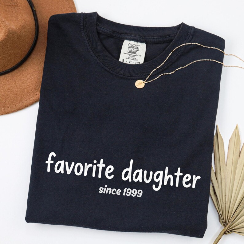 May include: A navy blue t-shirt with the words "favorite daughter since 1999" in white. A gold necklace with a small pendant rests on the shirt. A brown hat and dried palm leaf are in the background.