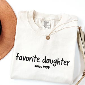 May include: Cream-coloured t-shirt with the words "favorite daughter" and "since 1999" printed in black. A gold necklace with a pendant and a brown hat are also visible.