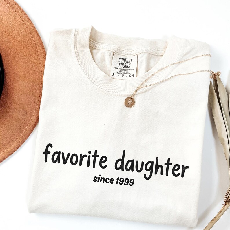 May include: Cream-coloured t-shirt with the words "favorite daughter" and "since 1999" printed in black. A gold necklace with a pendant and a brown hat are also visible.