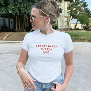以下が含まれることがあります： 胸元に赤い文字で「BAD DAY TO BE A HOT DOG」とプリントされた白い半袖Tシャツ。テキストの下中央には、黄色いマスタードがかかったホットドッグの小さなグラフィックイラストが描かれています。