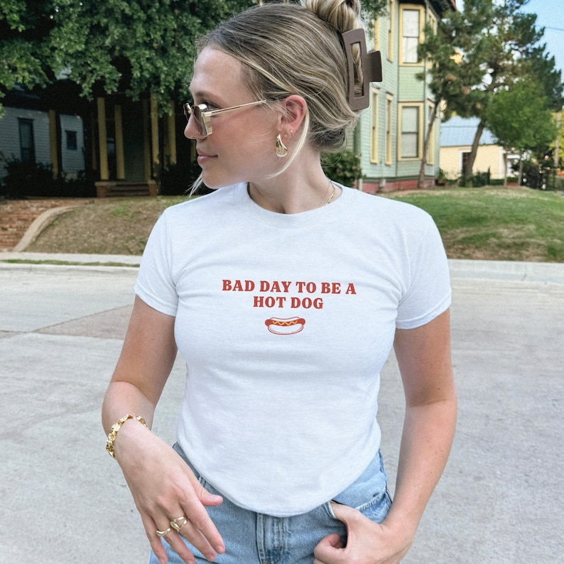 以下が含まれることがあります： 胸元に赤い文字で「BAD DAY TO BE A HOT DOG」とプリントされた白い半袖Tシャツ。テキストの下中央には、黄色いマスタードがかかったホットドッグの小さなグラフィックイラストが描かれています。