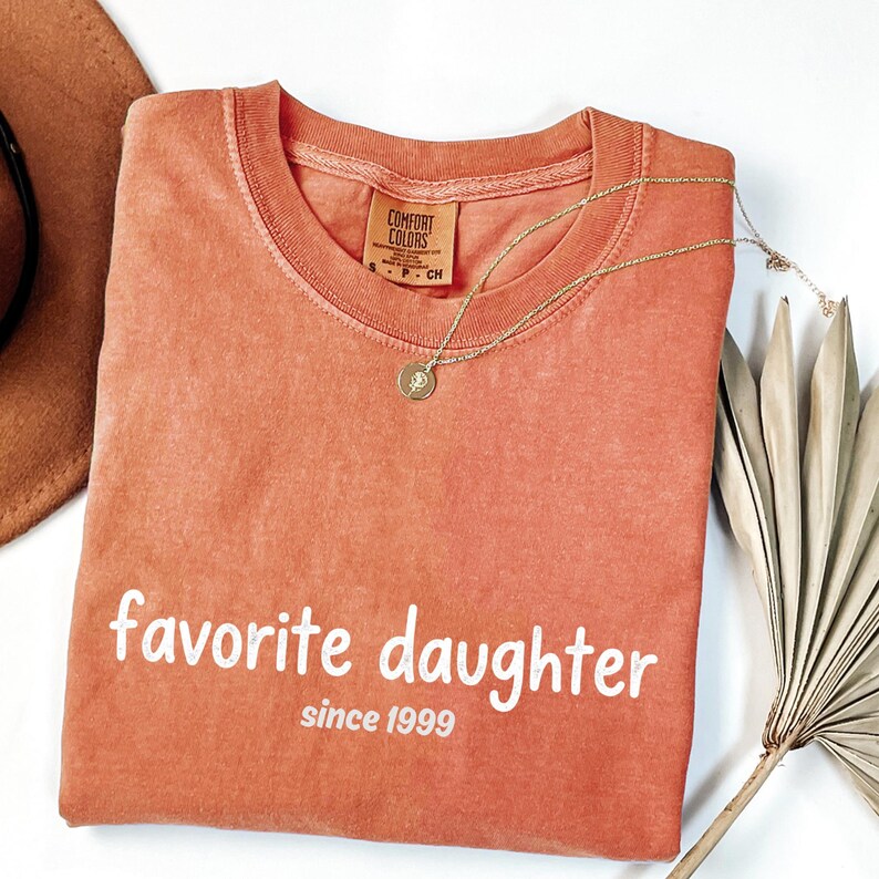 May include: Coral Comfort Colors t-shirt with the words "favorite daughter since 1999" printed in white. A gold necklace with a pendant rests on the shirt. A brown hat and dried palm leaf are in the background.