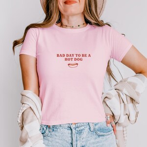 以下が含まれることがあります： 胸の中央に赤い文字で「BAD DAY TO BE A HOT DOG」とプリントされたライトピンクの半袖Tシャツ。文字の下には、マスタードのかかったホットドッグの小さなグラフィックイラストが描かれています。