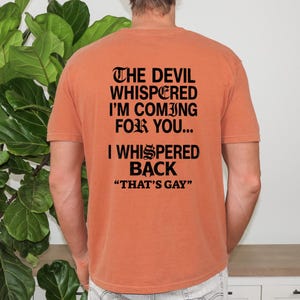 May include: A burnt orange short-sleeve t-shirt featuring black text on the back that reads: THE DEVIL WHISPERED I'M COMING FOR YOU... I WHISPERED BACK "THAT'S GAY". The shirt has a crew neckline and a standard fit, displaying the printed message clearly.