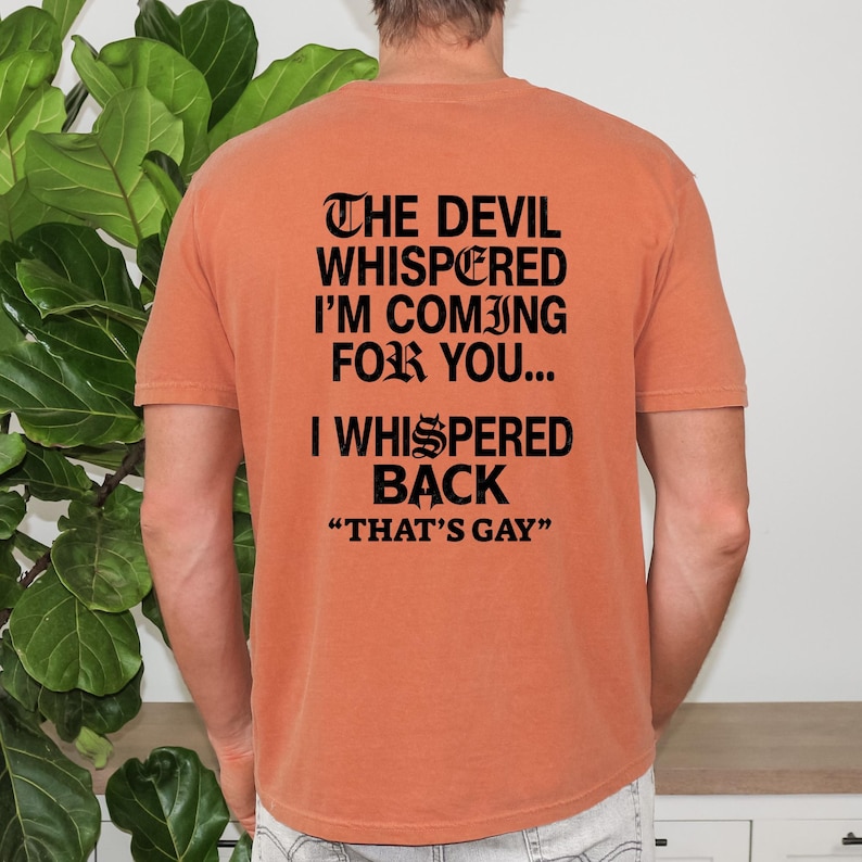 May include: A burnt orange short-sleeve t-shirt featuring black text on the back that reads: THE DEVIL WHISPERED I'M COMING FOR YOU... I WHISPERED BACK "THAT'S GAY". The shirt has a crew neckline and a standard fit, displaying the printed message clearly.