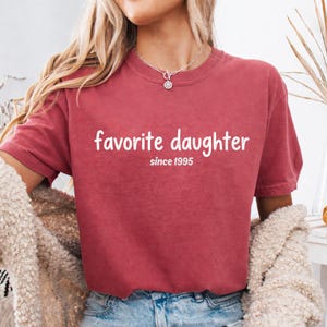May include: A maroon t-shirt with the words "favorite daughter" in white script, with "since 1995" below. The shirt has short sleeves and a crew neck. The person is wearing a necklace.