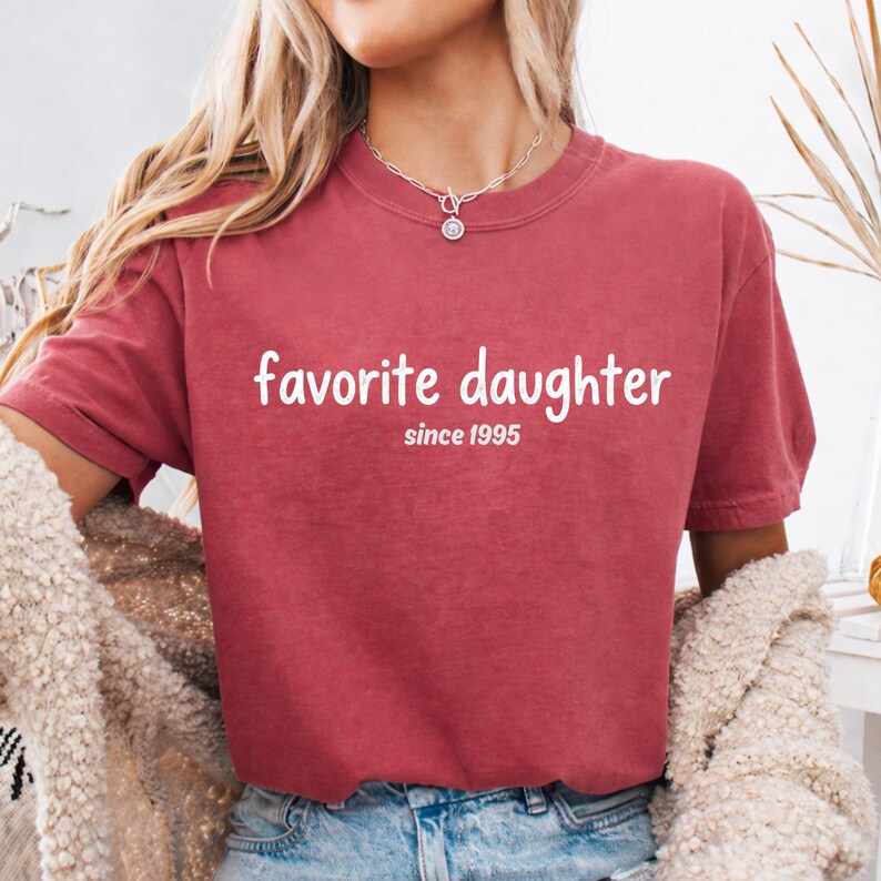 May include: A maroon t-shirt with the words "favorite daughter" in white script, with "since 1995" below. The shirt has short sleeves and a crew neck. The person is wearing a necklace.