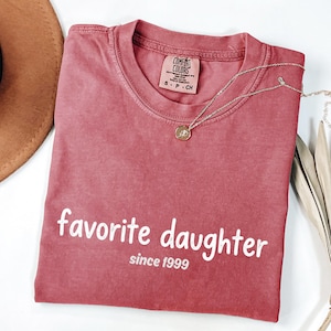 May include: A dusty rose Comfort Colors t-shirt with the words "favorite daughter" and "since 1999" printed in white. A silver necklace with a small pendant is draped across the shirt. The shirt is folded, and a brown hat is visible in the upper left corner.