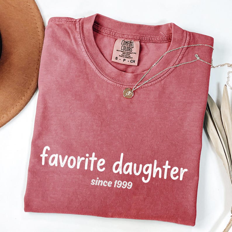 May include: A dusty rose Comfort Colors t-shirt with the words "favorite daughter" and "since 1999" printed in white. A silver necklace with a small pendant is draped across the shirt. The shirt is folded, and a brown hat is visible in the upper left corner.