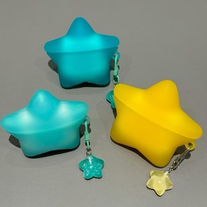May include: Three star-shaped containers in teal, yellow, and turquoise. Each container has a matching star charm attached by a small chain. The containers are made of a translucent material and sit on a gray surface.
