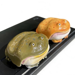 Puede incluir: Dos figuras de rana de cerámica. Una es de color verde oliva con manchas más oscuras, la otra es naranja con manchas rojas. Ambas tienen vientres blancos y bocas anchas y sonrientes. Las ranas están sobre una superficie negra.