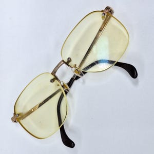 May include: Rimless spectacles with rectangular yellow-tinted lenses and gold-coloured metal frames. The temples are black and curve at the ends. The glasses are designed for vision correction.
