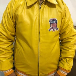 May include: A bright yellow leather jacket with a zip-up front and a collared neckline. The jacket features a patch on the chest with the text "AVIREX ESTABLISHED 1975". The cuffs and hem have orange and white striped detailing.