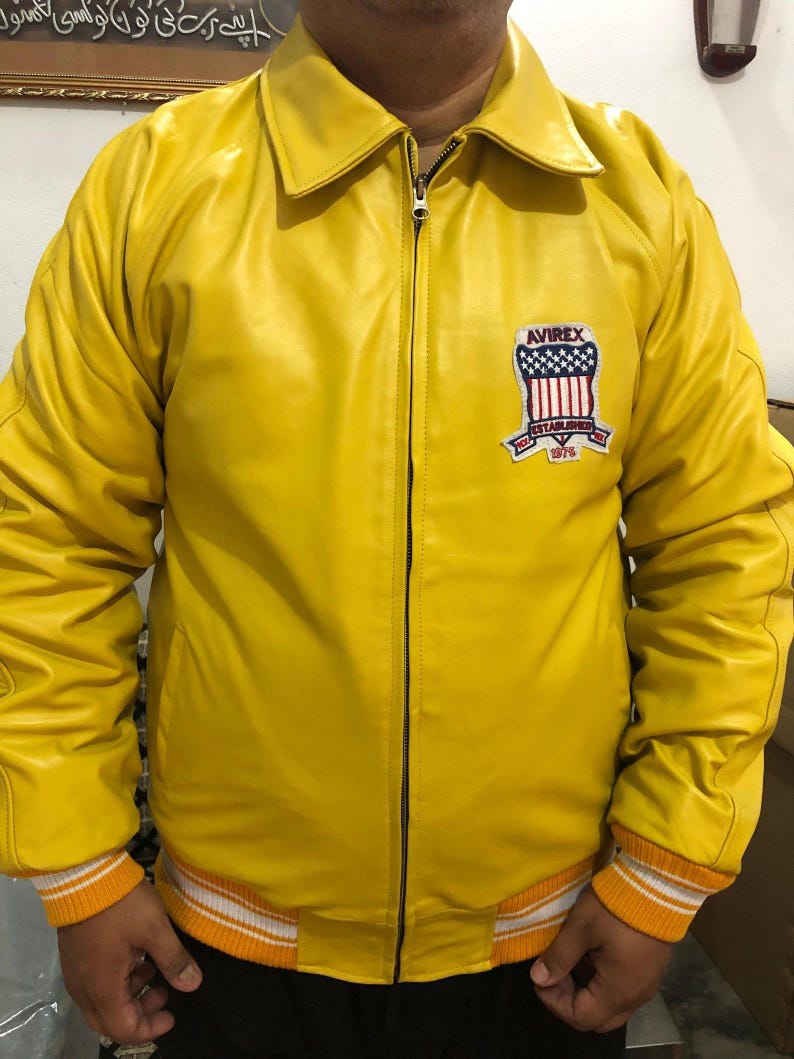 May include: A bright yellow leather jacket with a zip-up front and a collared neckline. The jacket features a patch on the chest with the text "AVIREX ESTABLISHED 1975". The cuffs and hem have orange and white striped detailing.
