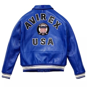 May include: A vibrant blue leather jacket featuring the word "AVIREX" in white across the upper back. Below, the word "USA" is displayed in white. A circular patch with the words "ALL AMERICANS" and "ESTABLISHED 1975" is centered above the "USA" text.