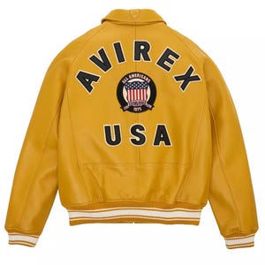 May include: Mustard yellow leather jacket with the word "AVIREX" arched across the shoulders. A circular patch with the American flag and the words "ALL AMERICANS ESTABLISHED 1975" is centered below the word "AVIREX". The word "USA" is printed below the patch.