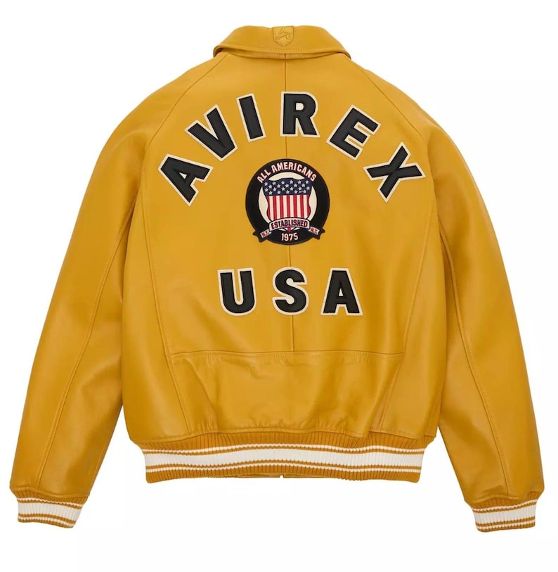 May include: Mustard yellow leather jacket with the word "AVIREX" arched across the shoulders. A circular patch with the American flag and the words "ALL AMERICANS ESTABLISHED 1975" is centered below the word "AVIREX". The word "USA" is printed below the patch.