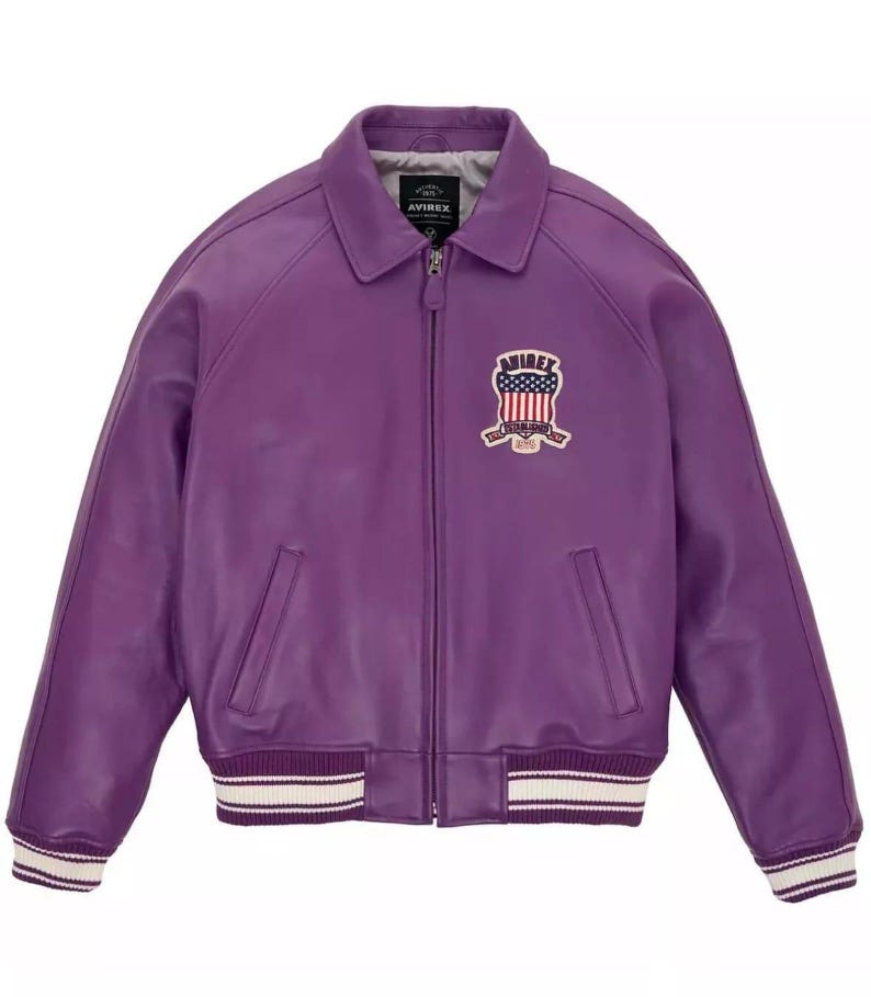 K&ouml;nnte beinhalten: Eine lila Lederjacke mit Rei&szlig;verschluss, Kragen und zwei Vordertaschen. Die Jacke hat einen Aufn&auml;her auf der Brust mit einem amerikanischen Flaggenmuster und dem Wort "AVIREX". Die Manschetten und der Saum haben wei&szlig;e und cremefarbene Streifen.