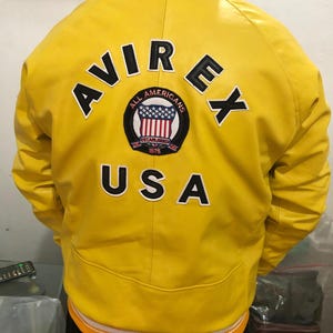 May include: A bright yellow leather jacket with the word "AVIREX" and "USA" in black lettering. The jacket features a patch with the words "ALL AMERICANS" and the American flag. The jacket has an orange and white striped trim.