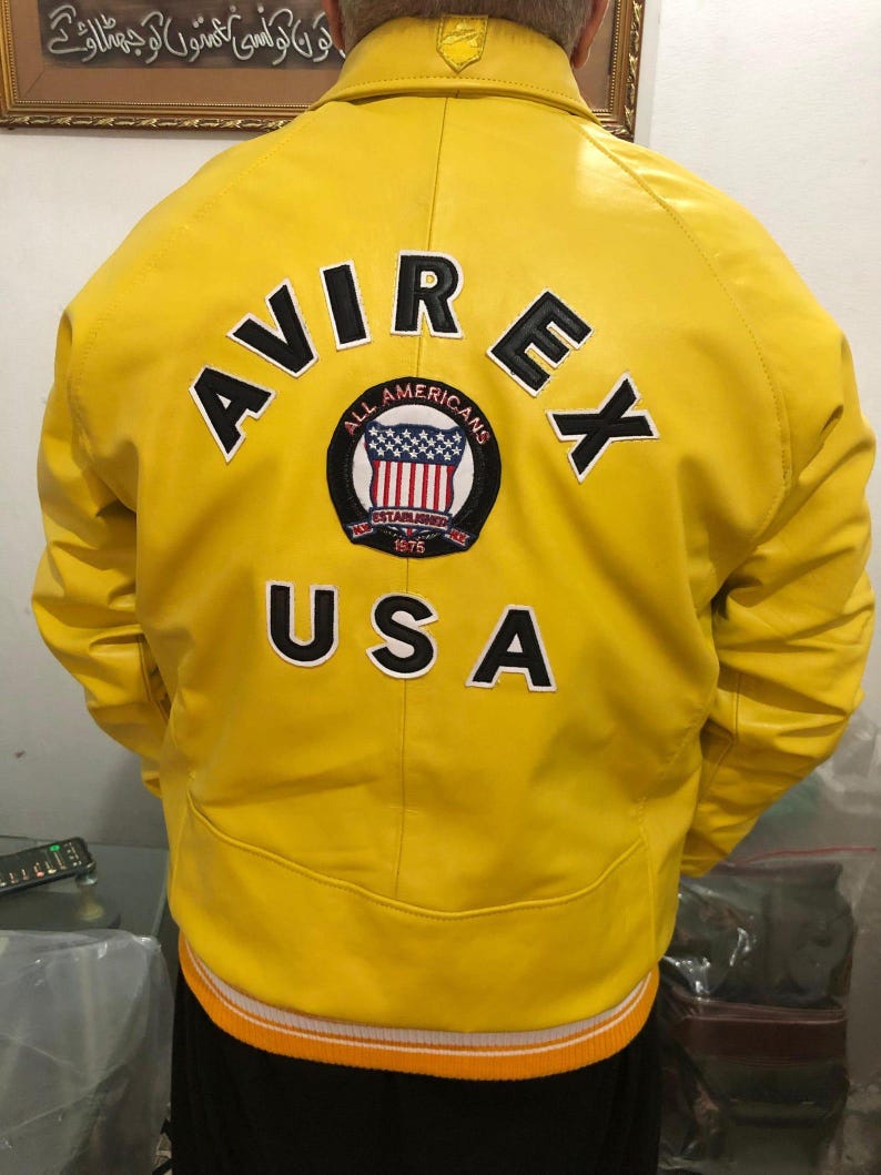 May include: A bright yellow leather jacket with the word "AVIREX" and "USA" in black lettering. The jacket features a patch with the words "ALL AMERICANS" and the American flag. The jacket has an orange and white striped trim.