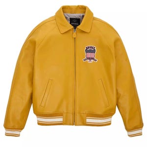 May include: Mustard yellow leather jacket with a collared neckline and a zipper closure. Features a patch with a red, white, and blue design on the chest. The cuffs and hem have white stripes. The brand name "AVIREX" is visible.