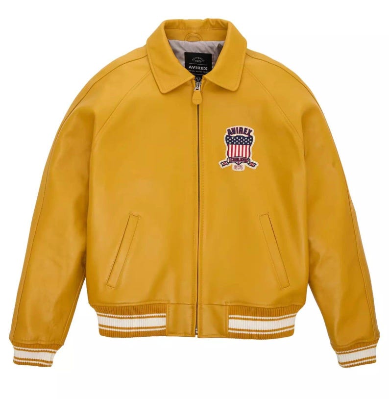 May include: Mustard yellow leather jacket with a collared neckline and a zipper closure. Features a patch with a red, white, and blue design on the chest. The cuffs and hem have white stripes. The brand name "AVIREX" is visible.