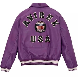 K&ouml;nnte beinhalten: Eine lila Lederjacke mit dem Wort "AVIREX" auf dem oberen R&uuml;cken und "USA" unter einem kreisf&ouml;rmigen Aufn&auml;her mit einem amerikanischen Flaggen-Design. Die Jacke hat einen Kragen, gerippte Manschetten und einen wei&szlig;-schwarz gestreiften Saum.