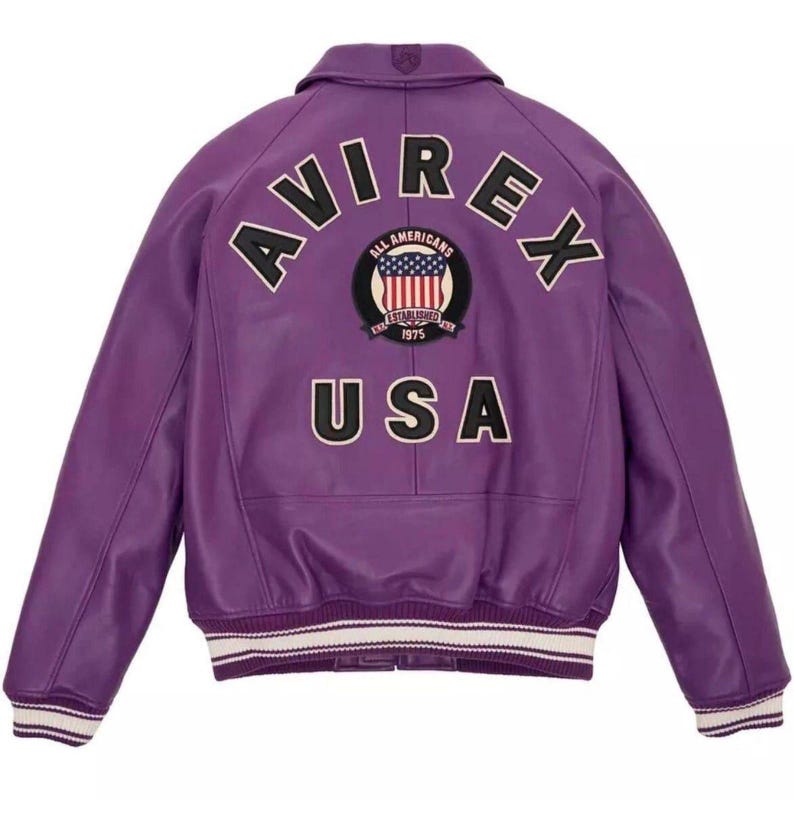 K&ouml;nnte beinhalten: Eine lila Lederjacke mit dem Wort "AVIREX" auf dem oberen R&uuml;cken und "USA" unter einem kreisf&ouml;rmigen Aufn&auml;her mit einem amerikanischen Flaggen-Design. Die Jacke hat einen Kragen, gerippte Manschetten und einen wei&szlig;-schwarz gestreiften Saum.